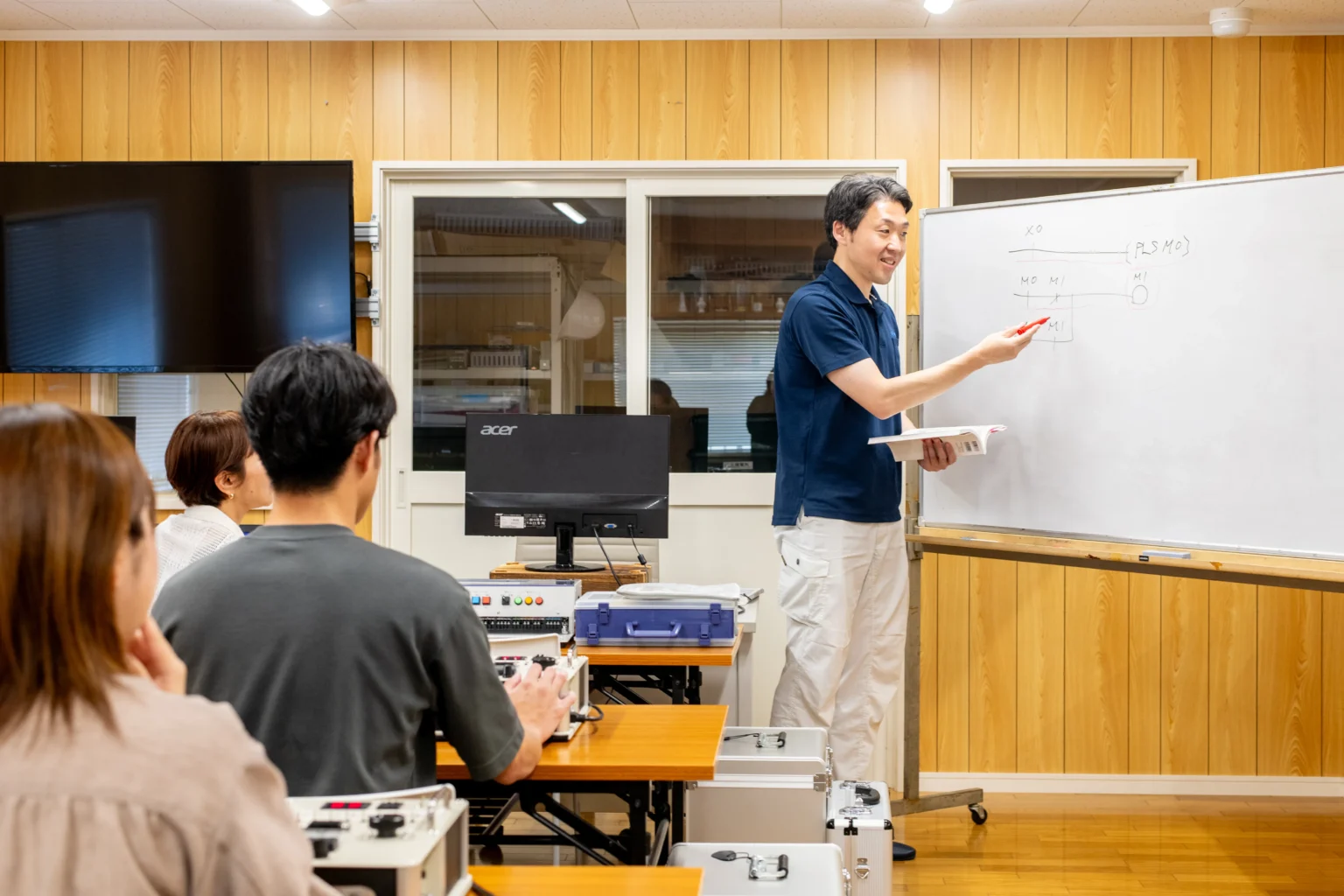 エンジニア向け技術研修スクールで講師がホワイトボードを使って回路設計を解説し実技装置を備えたデスクで受講生が熱心に学ぶ