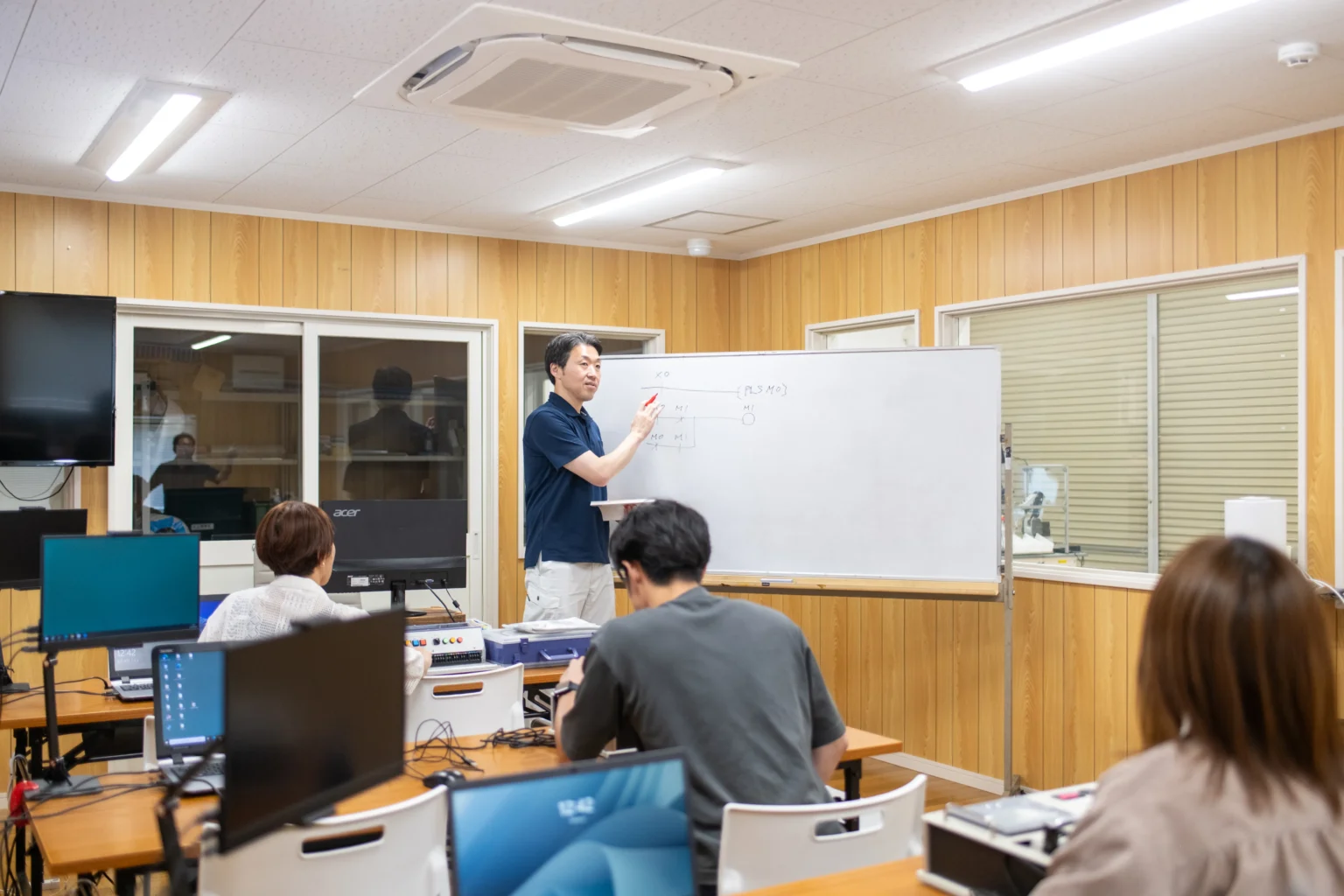 エンジニアスクールの技術研修にて専門講師がホワイトボードでシーケンス制御回路を解説しPC操作する受講生に実践指導する