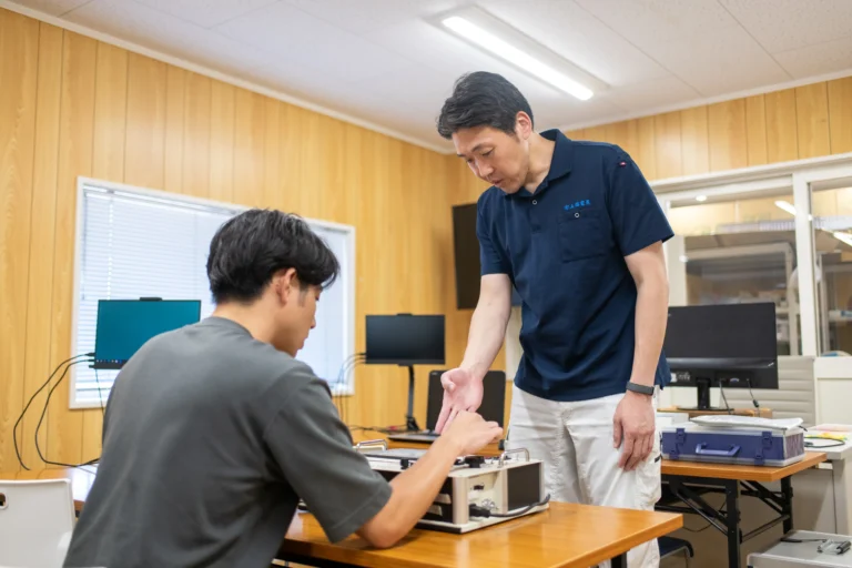 エンジニア研修にて熟練指導員が実技装置を操作する受講生に寄り添い実践的な技術ノウハウや機械制御のスキルを丁寧に教える