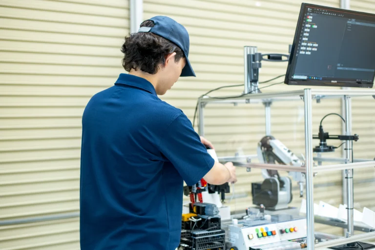 研修室で機械制御を行うエンジニアがモニターに表示されたプログラムを確認しつつロボットアームや機械装置の調整学習に取り組む