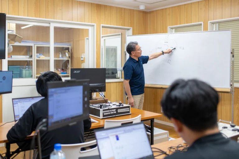 技術研修スクールにて専門講師がホワイトボードを使って工程図を解説しパソコンで制御学習に取り組むエンジニア受講生に指導する