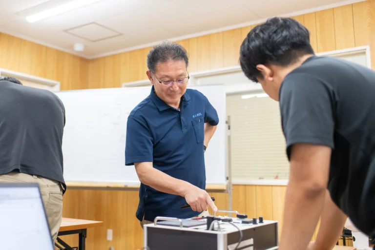 技術研修やエンジニア教育の現場で経験豊富な指導員が機械制御装置を手元で指さしながら若手受講生に実践的なスキルを分かりやすく解説する