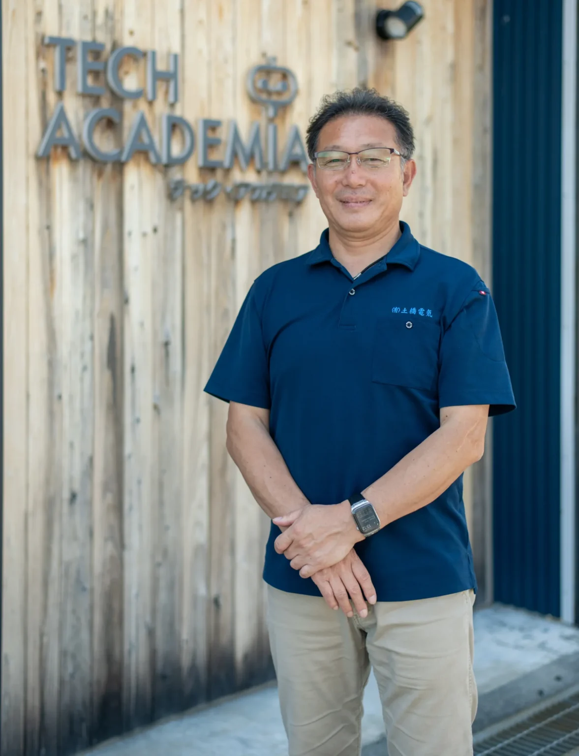 エンジニア向けスクールTECH ACADEMIAで指導を行う講師が木目調の施設前に立ち温かい笑顔で受講生を迎え入れる信頼感あふれる姿