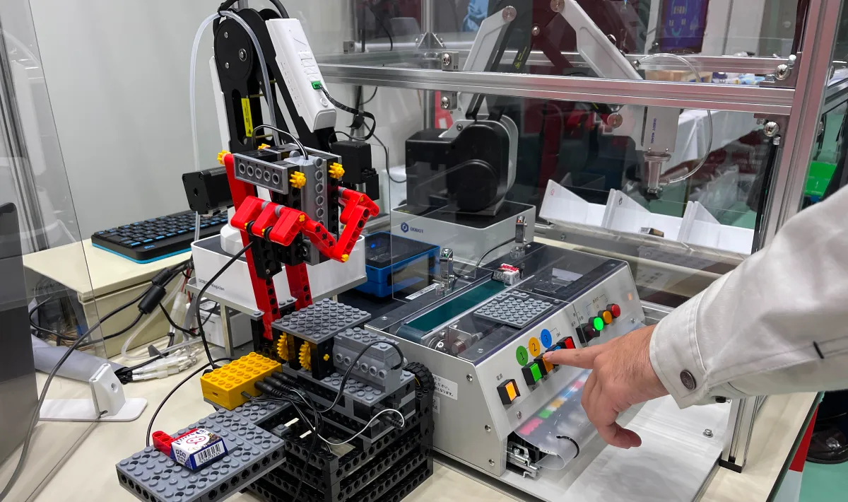 作業着姿のエンジニアがブロックで構築された小型ロボットアームとコンベアを備えた自動制御システムの操作盤で青いボタンを押し機器制御の開発テストを行っている