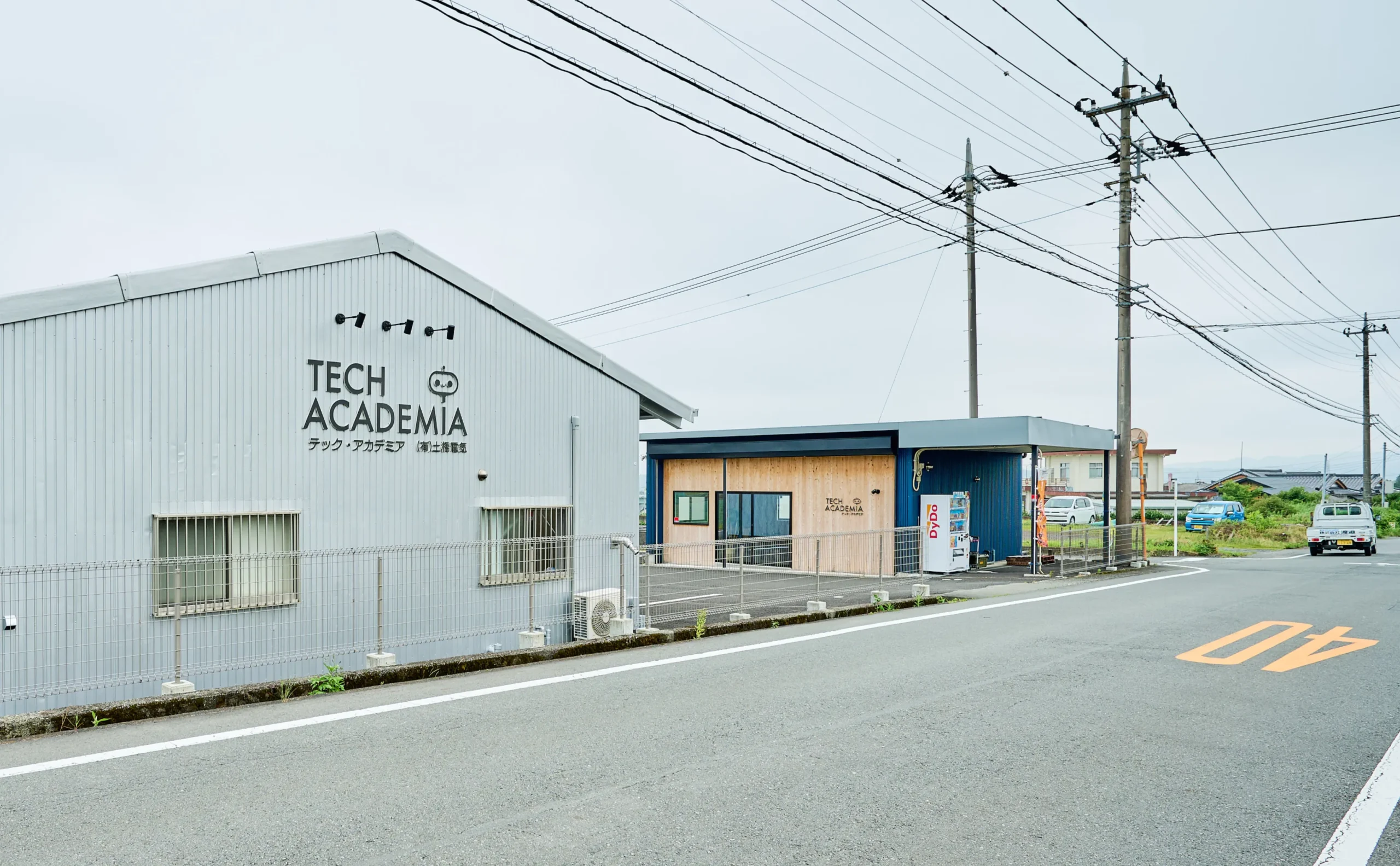 曇り空の下にあるTECH ACADEMIAの看板を掲げた薄青い波板の外壁の建物と隣接する木目調の建物の前に舗装された道路が広がる静かな風景