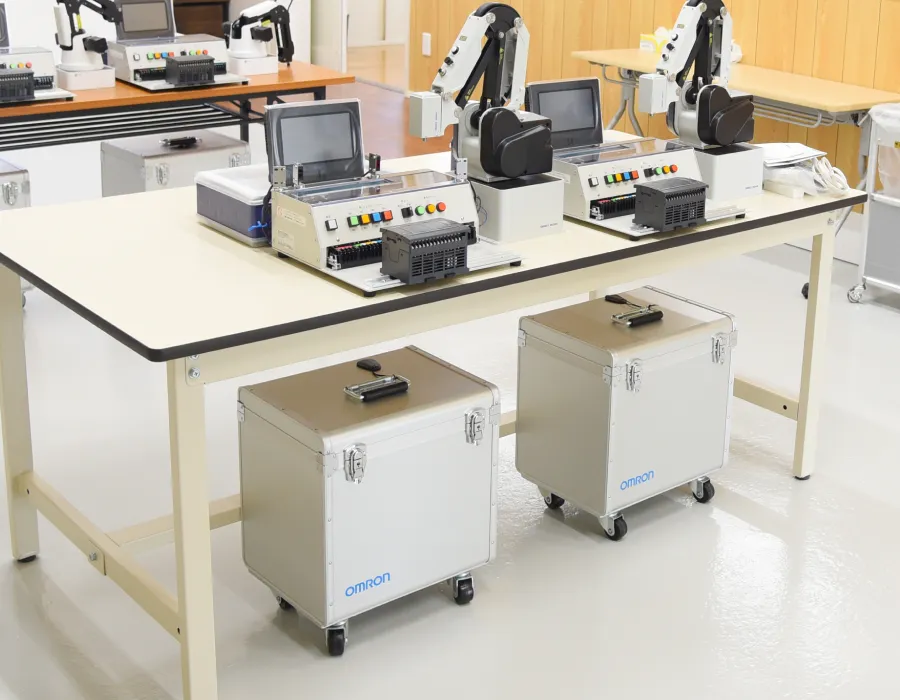 制御機器や白いロボットアームが長机に複数並べられ工場の自動化技術を実践的に学べる明るく整備されたエンジニア向け実習エリア