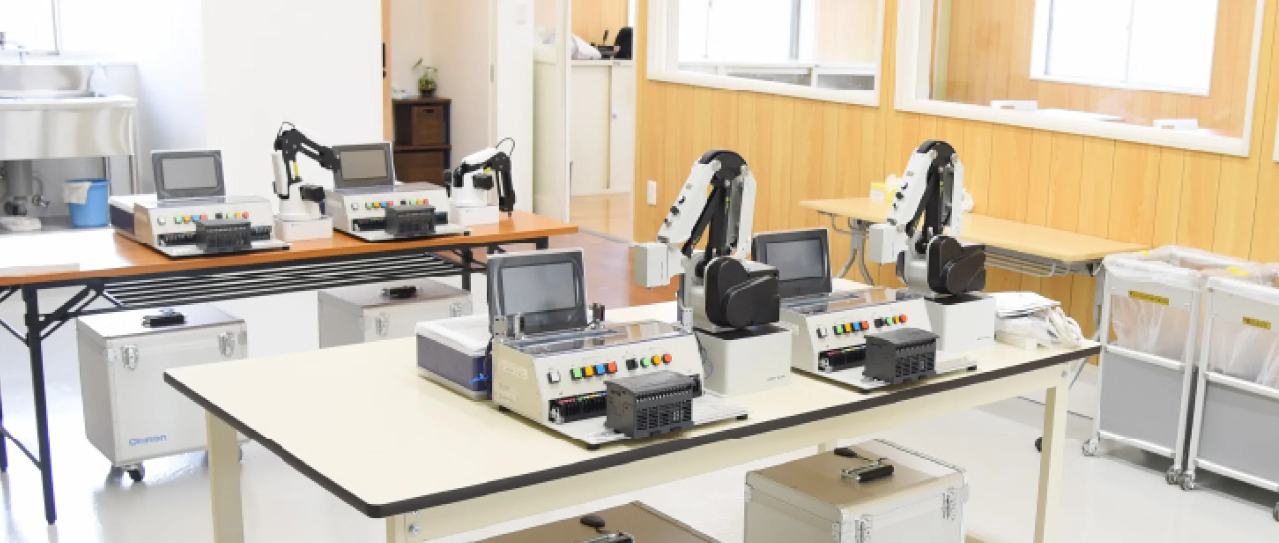 制御機器や白いロボットアームが長机に複数並べられ工場の自動化技術を実践的に学べる明るく整備されたエンジニア向け実習エリア